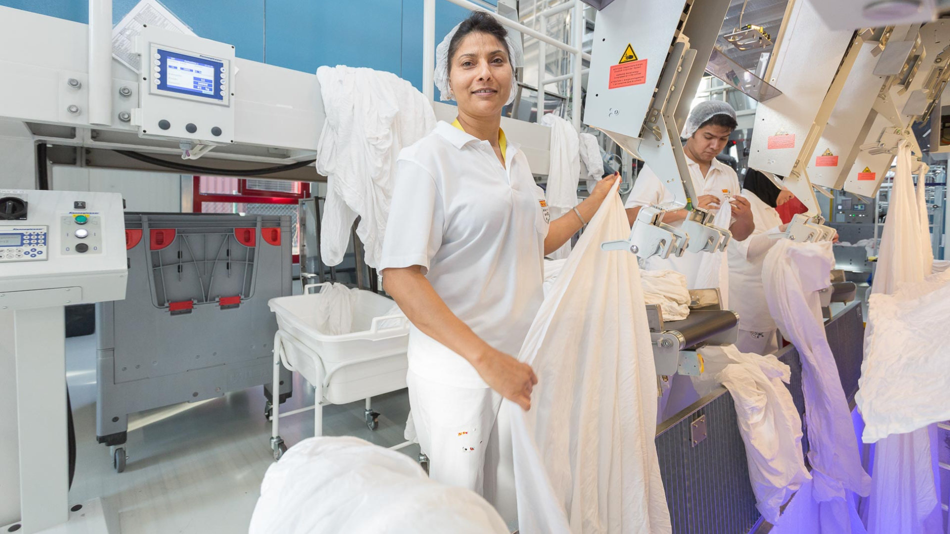 Employees in an industrial laundry folding freshly cleaned bed linen at modern machines.