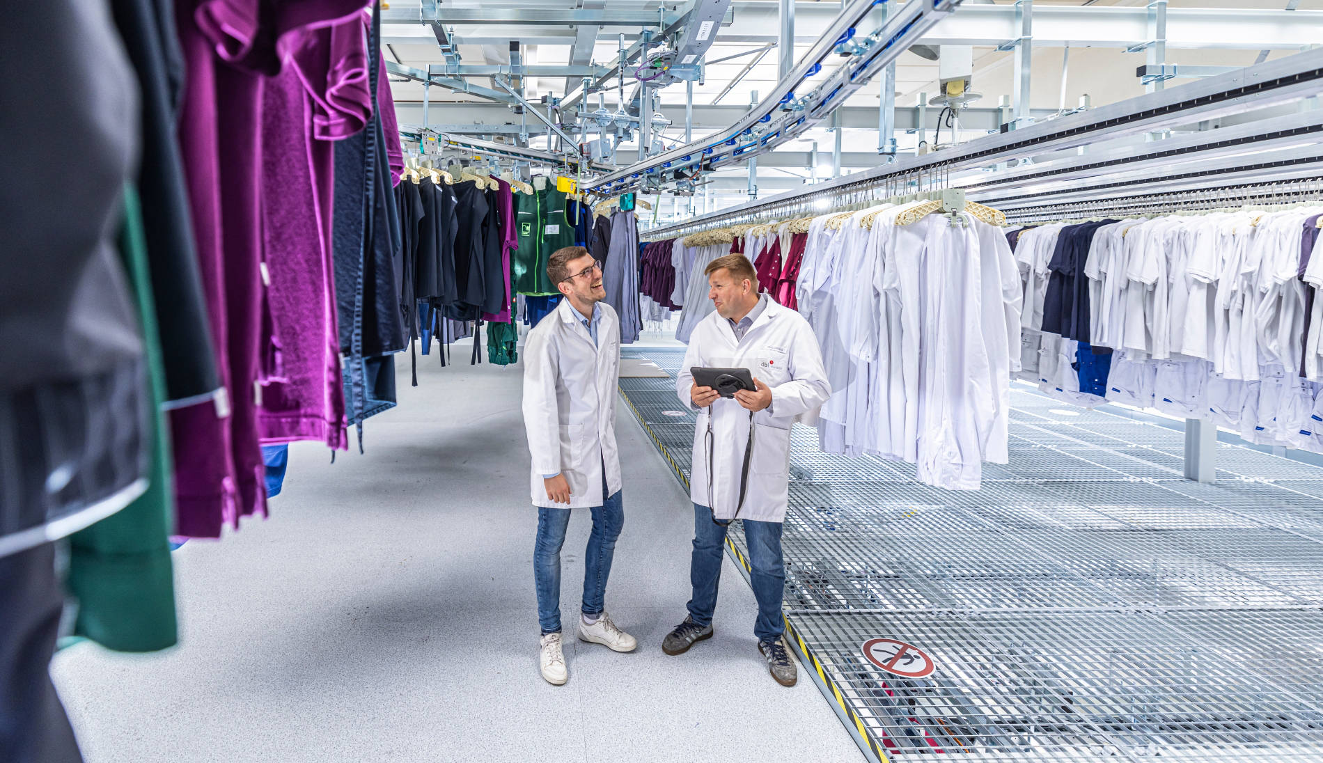Zwei Mitarbeiter in weißer Arbeitskleidung besprechen Prozesse in automatisierter Textilreinigungsanlage mit hängender Kleidung.